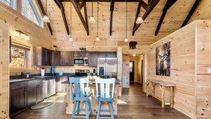 A modern kitchen with wooden cabinetry, stainless steel appliances, and a dining area with two blue chairs. - Peaceful Mountain Escape with Ridge Views & Hot Tub (Maggie Valley)