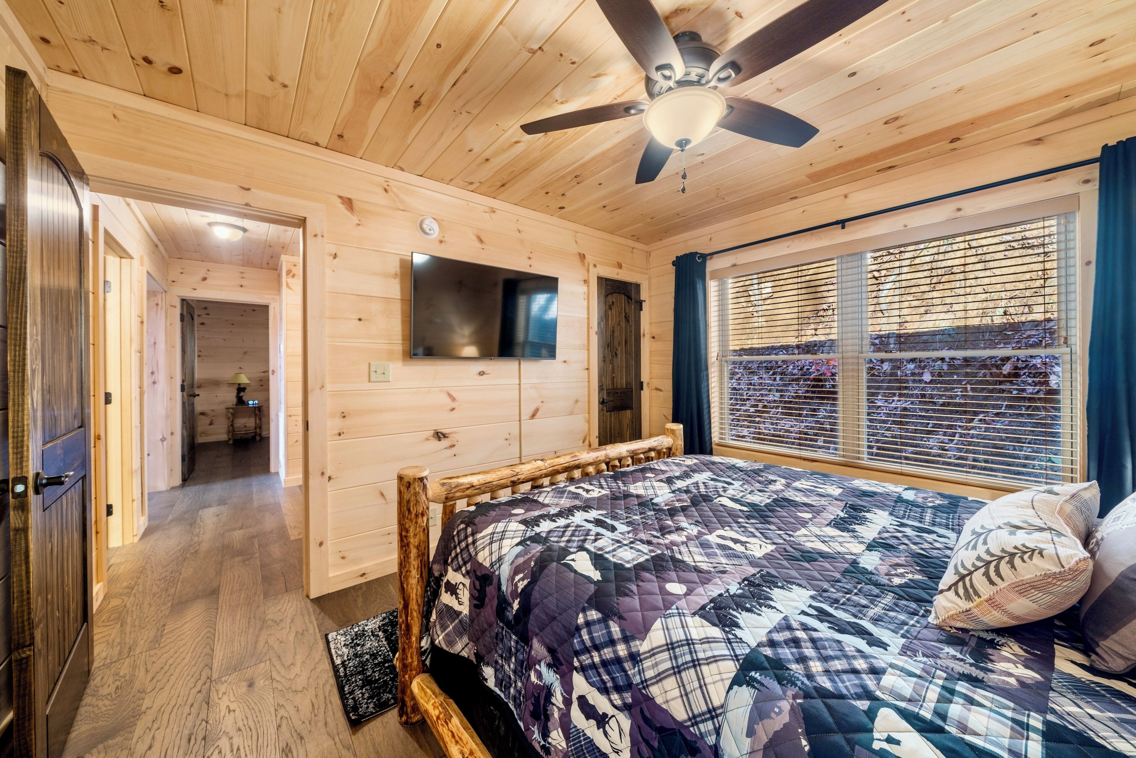 Un dormitorio de cabaña de madera con cama, ventilador de techo, televisor montado y ventana con persianas.