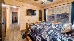 A wooden cabin bedroom with a bed, a ceiling fan, a mounted TV, and a window with blinds. - Peaceful Mountain Escape with Ridge Views & Hot Tub (Maggie Valley)