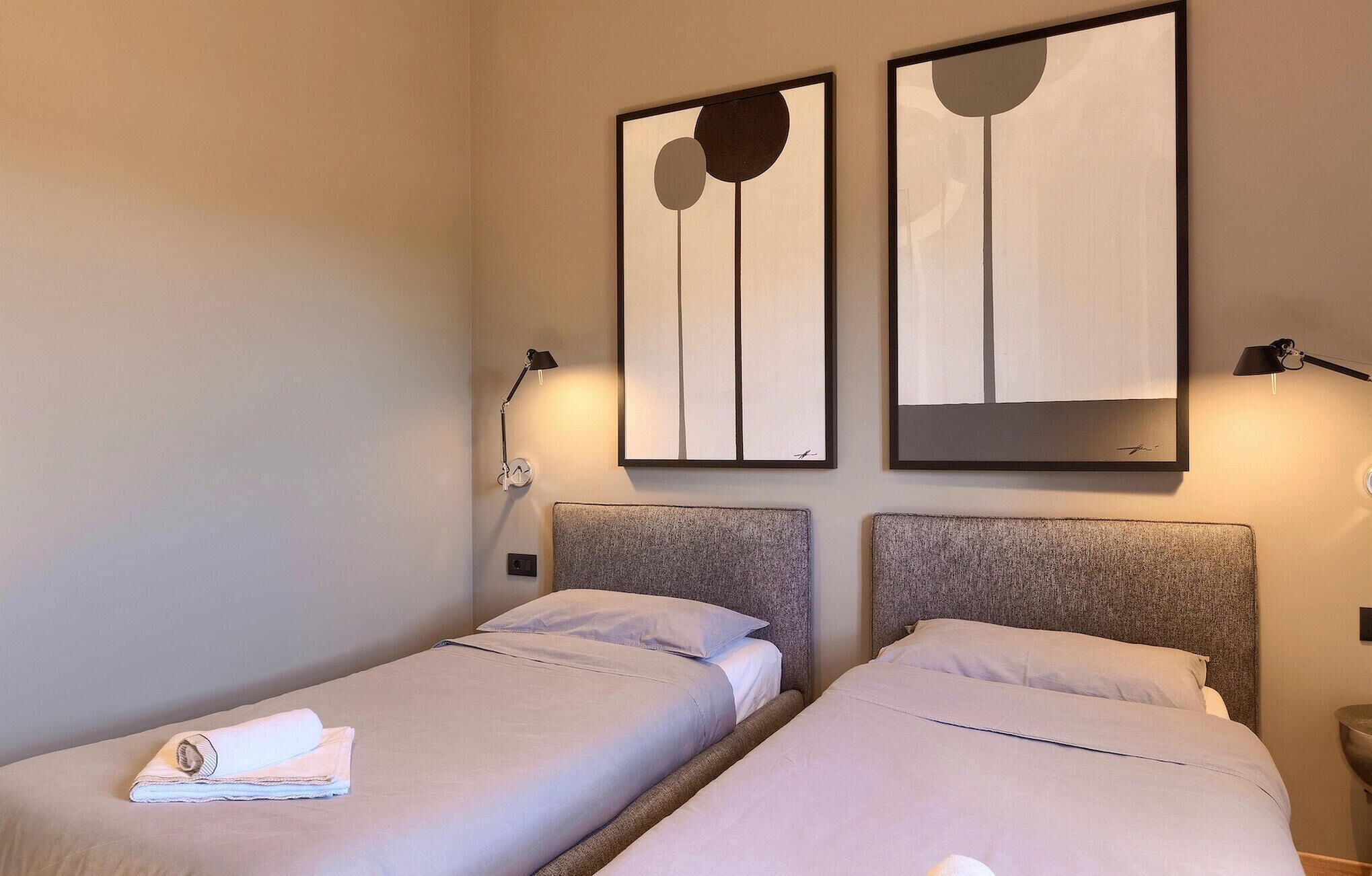 A hotel room with two beds, each with a headboard, a wall-mounted artwork, a bedside lamp, and a folded towel on one bed.