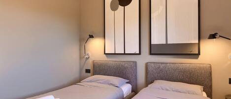 A hotel room with two beds, each with a headboard, a wall-mounted artwork, a bedside lamp, and a folded towel on one bed.