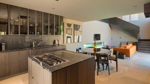 A modern kitchen with dark wood cabinets, a marble countertop, and a central island with a stove. There is a dining area with a round table and chairs, and a living space with a sofa and a TV.