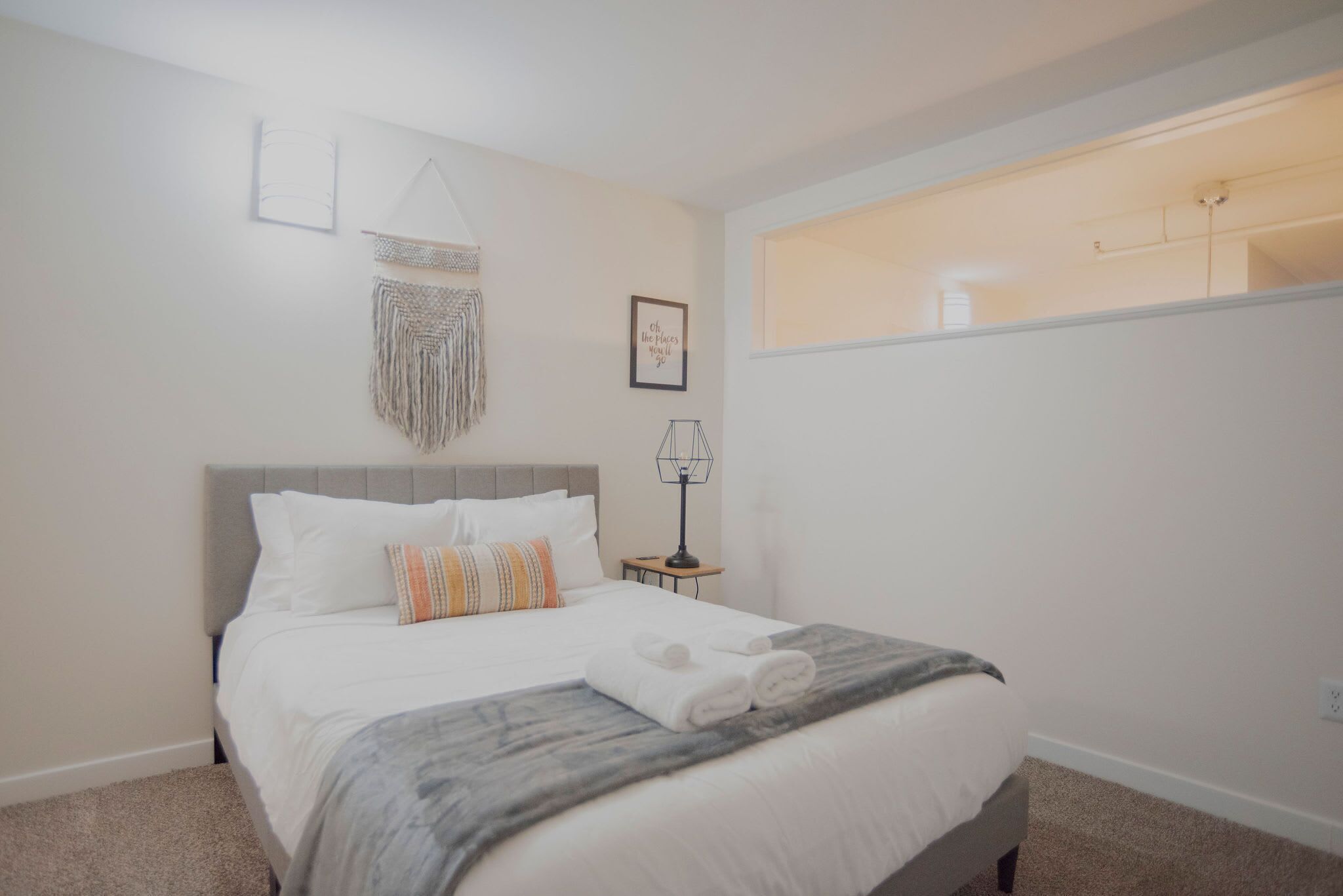 A neatly arranged bedroom with a bed, a bedside table, a wall-mounted lamp, and a decorative macrame piece.