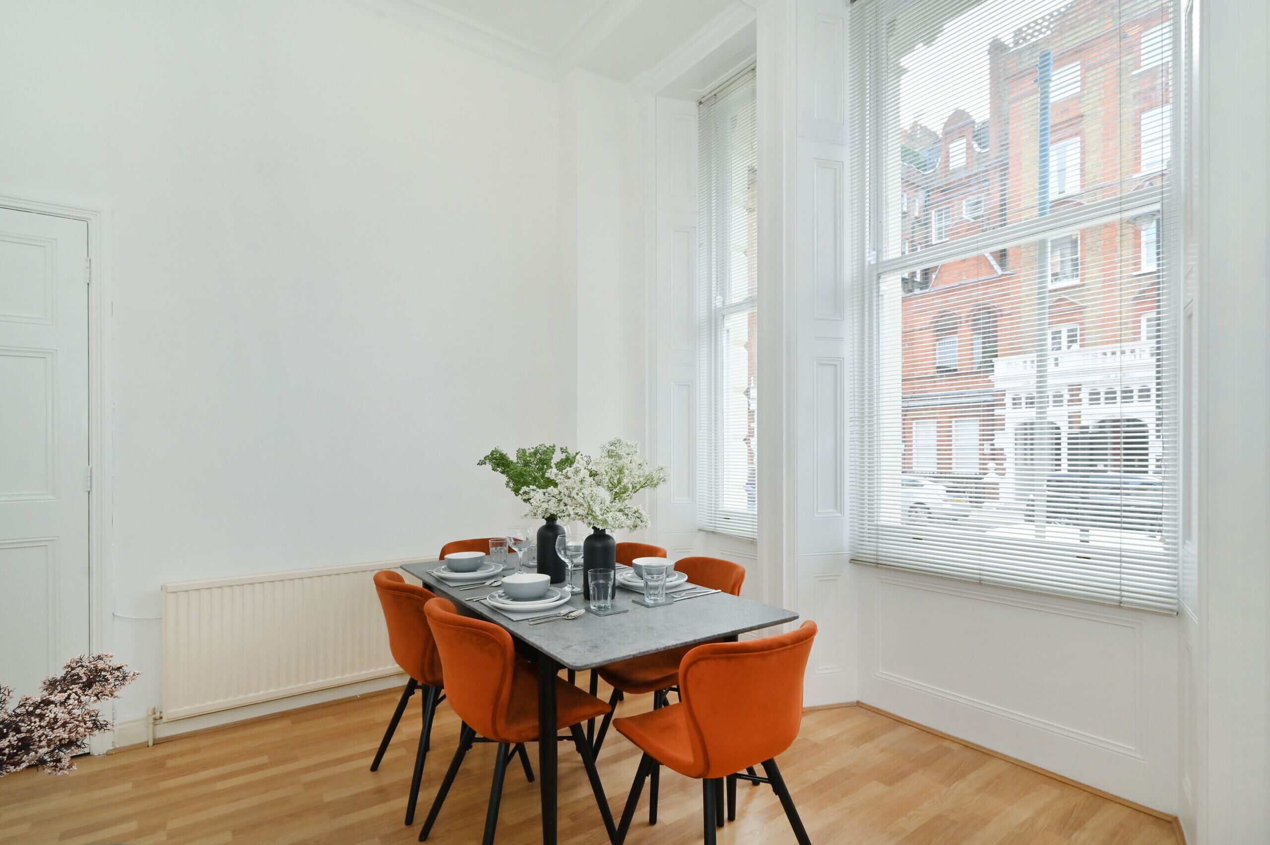 A dining area with a table set for four, surrounded by orange chairs, white walls, and large windows with blinds.