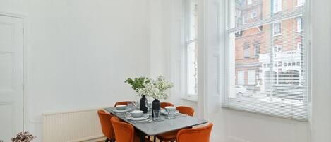 A dining area with a table set for four, surrounded by orange chairs, white walls, and large windows with blinds.