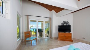 Hotelkamer met een bed, een televisie, een houten dressoir, een stoel en een schuifdeur naar een balkon met uitzicht op de oceaan.