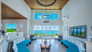 Een ruime woonkamer met een prachtig uitzicht op de oceaan, twee blauwe banken met kussens met sterrenmotief, een houten salontafel en een moderne kroonluchter.