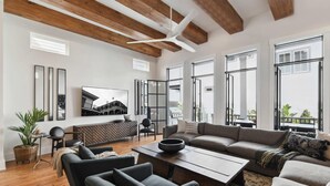 A spacious living room with a large sectional sofa, a coffee table, and a TV mounted on the wall. There are large windows providing natural light and a view of the outside. - New Luxury Home | Heated Pool & Spa (New Orleans)