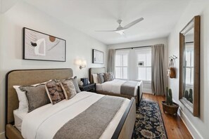 Habitación de hotel con dos camas, un ventilador de techo, un ventanal con cortinas, una lámpara de pared y un cuadro enmarcado.