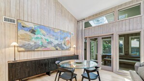 Ein moderner Essbereich mit Glastisch und Stühlen, einem großen abstrakten Gemälde an der Wand und Blick durch Glastüren.