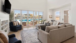 A spacious living room with a large sofa, a smaller armchair, and a flat-screen TV mounted on the wall. The room has large windows offering a view of a pool and buildings. - Experience Utah at the Beach: Desert Color Lagoon + Beaches (St. George)