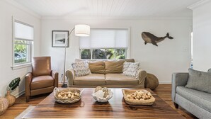 A living room with a brown leather armchair, a beige sofa with patterned cushions, a wooden coffee table with baskets of potatoes, and a whale wall decoration.