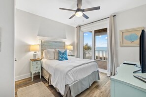 Quarto com cama, mesa de cabeceira, abajur, ventilador de teto e vista para o mar através de uma porta de correr.