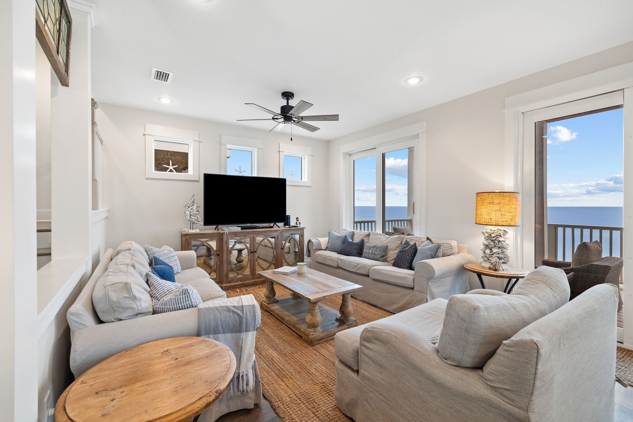 A spacious living room with a ceiling fan, large windows, a flat-screen TV, and a view of the ocean.