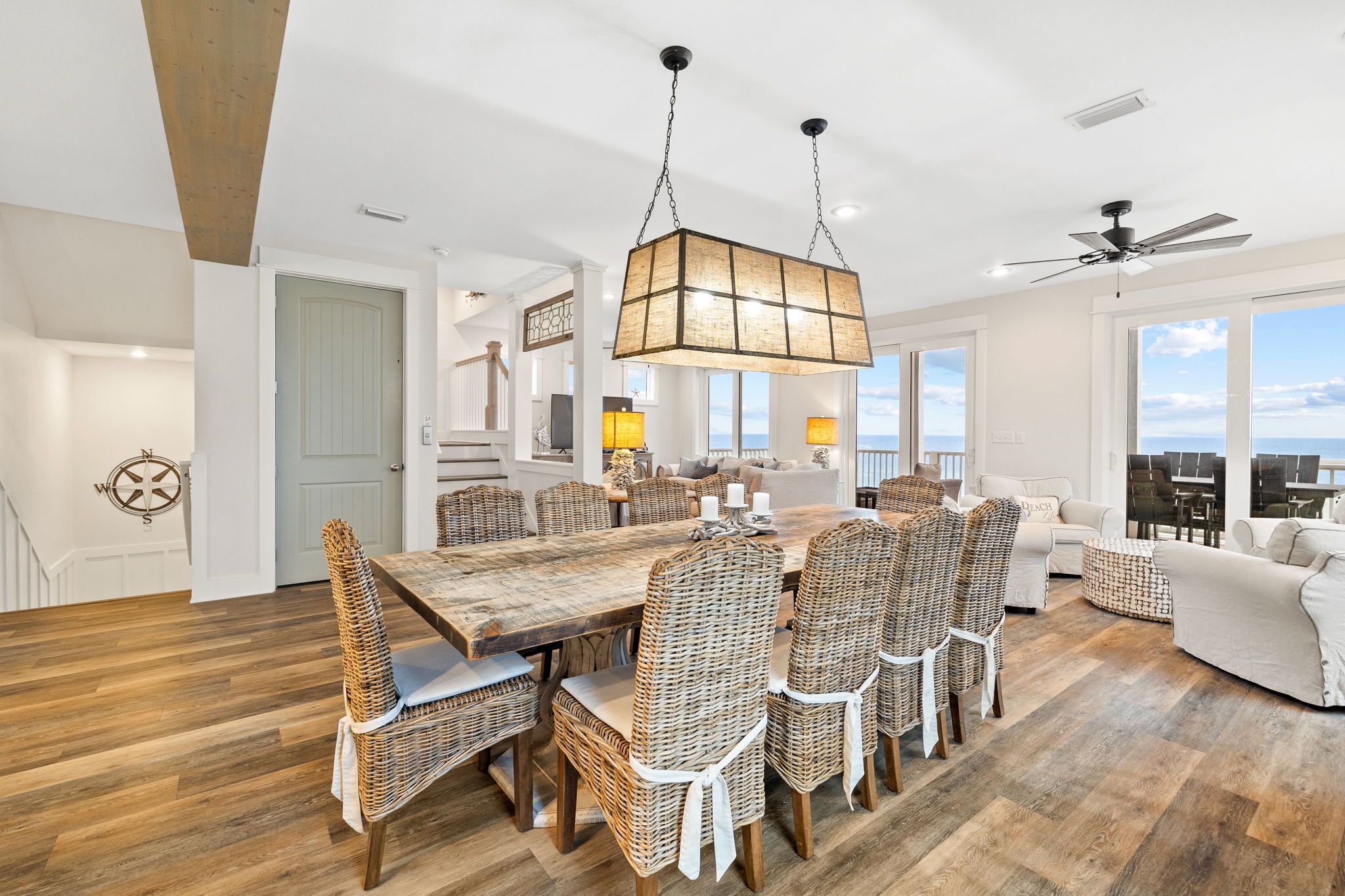A spacious living room with a large dining table, wicker chairs, a ceiling fan, and large windows offering a view.