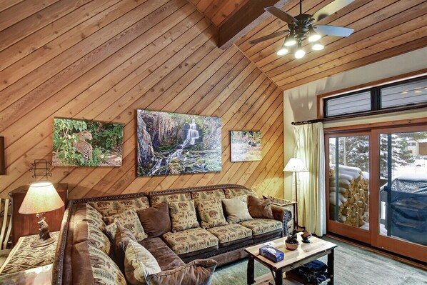A cozy living room with a wooden ceiling, a sofa with patterned cushions, a coffee table, and a large window with a view of the outside. - Snowcreek 464- Nice Condo with Mountain Views (Mammoth Lakes)