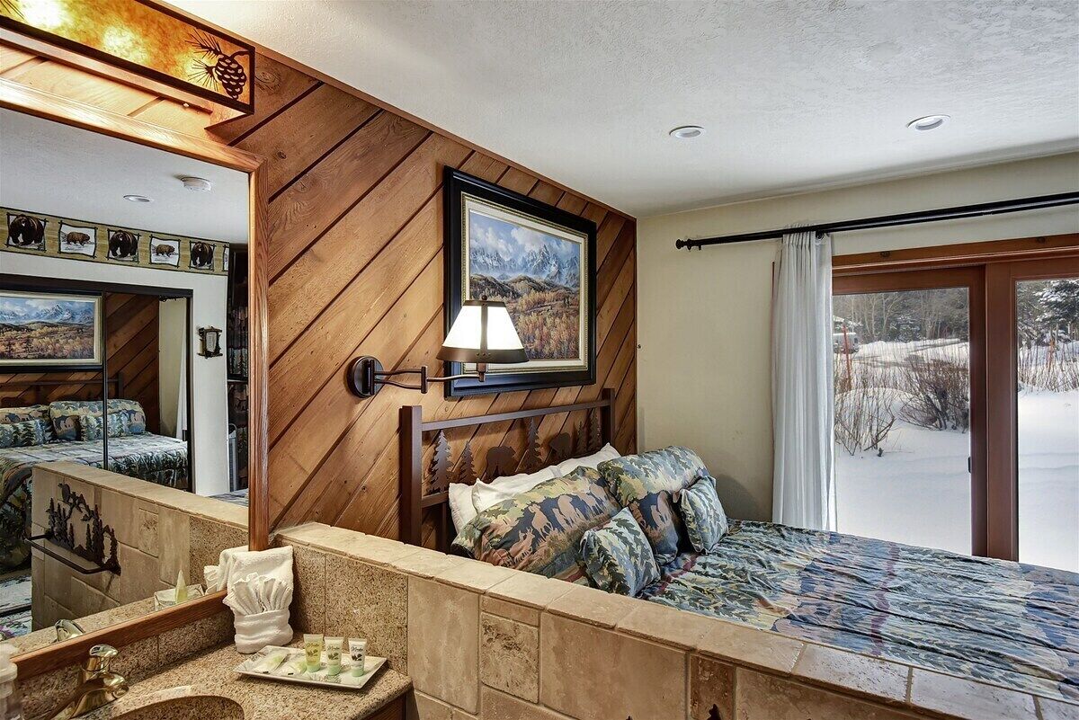 Une chambre à coucher avec une tête de lit en bois, un lit recouvert d’une couette à motifs camouflage, une table de chevet avec une lampe, un miroir et une fenêtre donnant sur un paysage enneigé.