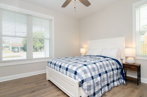 A bedroom with a bed, a ceiling fan, a nightstand with a lamp, and large windows with blinds.
