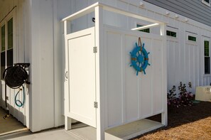 A white utility shed with a blue ship's wheel decoration, a hose, and a small garden with pink flowers.