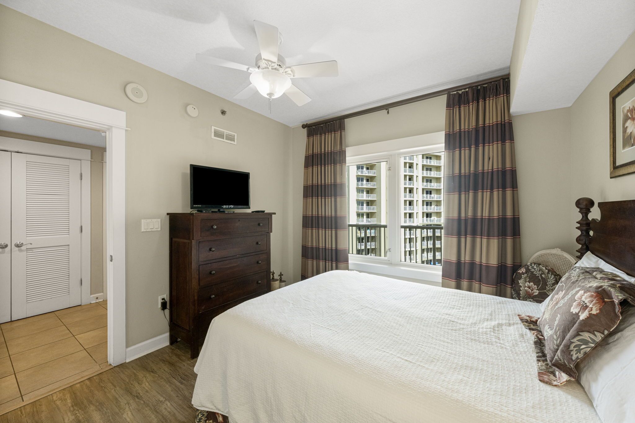 Une chambre à coucher comprenant un lit, une commode, un téléviseur, un ventilateur de plafond et une fenêtre avec des rideaux.