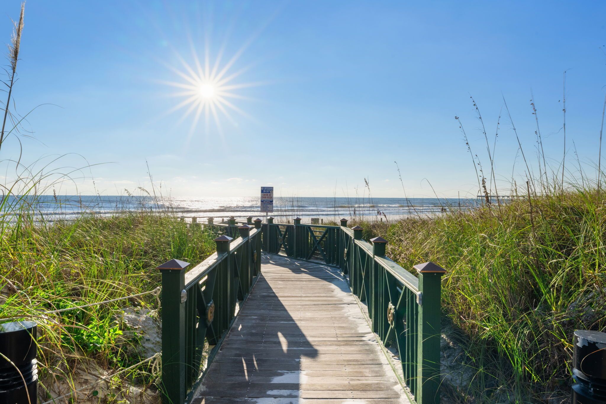A wooden boardwalk leading to a beach with a clear sky and the sun shining.