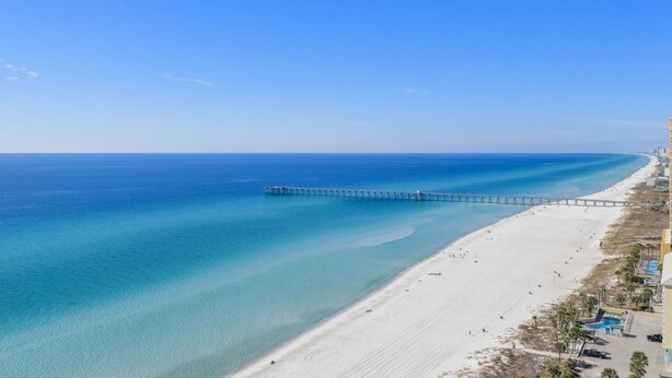 A beach with a pier extending into the ocean, a sandy shore, and a building with a pool on the right.