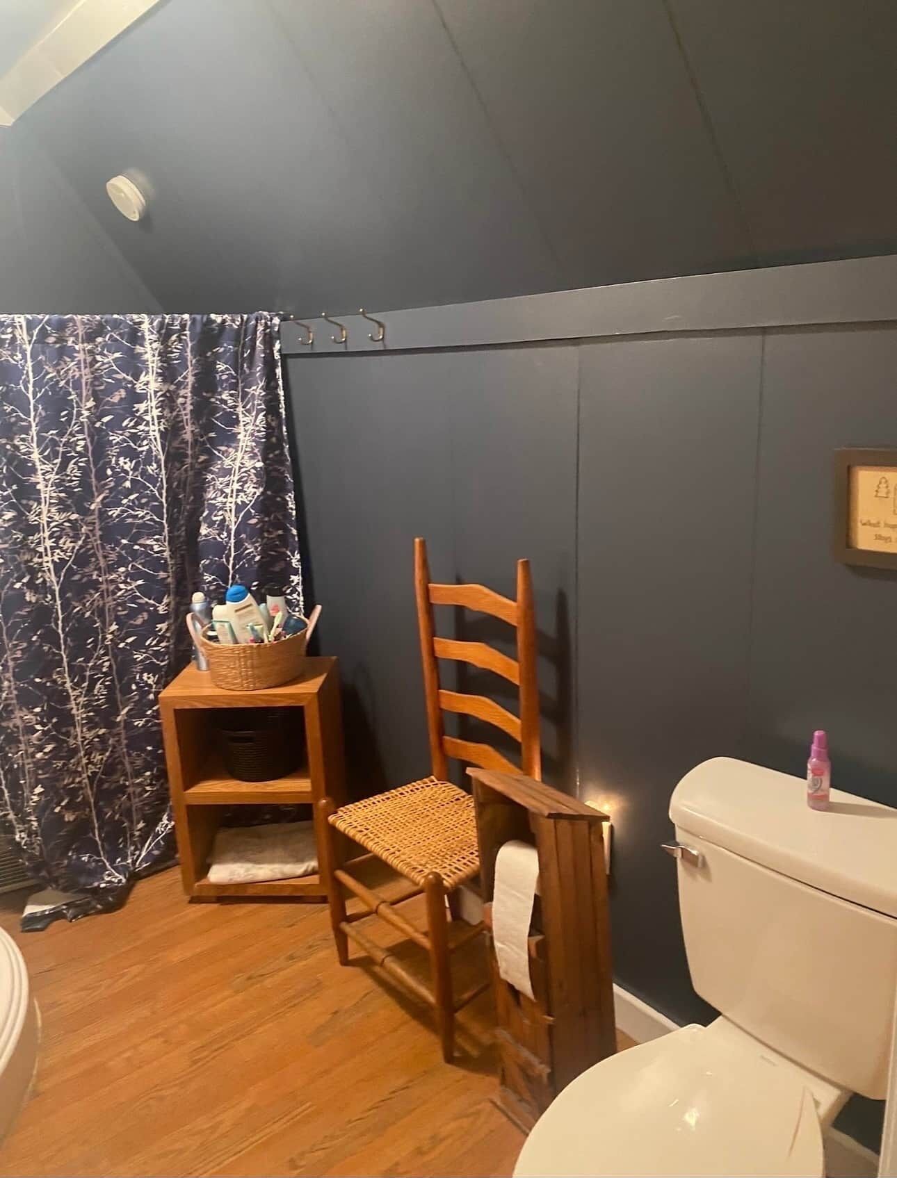 Un baño con una silla de madera, un estante pequeño de madera con artículos de aseo, un inodoro y una pared oscura.