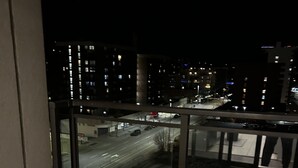 Vista nocturna desde un balcón con barandilla de vidrio que da a una calle y edificios de la ciudad.