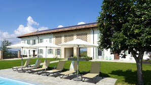 A poolside area with lounge chairs and umbrellas, a stone pool, and a building with a tiled roof and shutters. - Luxury home with pool and spectecular views (Piemonte)