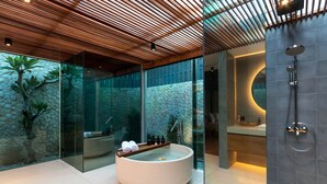 A modern bathroom with a large freestanding bathtub, a glass-enclosed shower area, and a stone wall backdrop. - Navya Villas (Kerobokan)