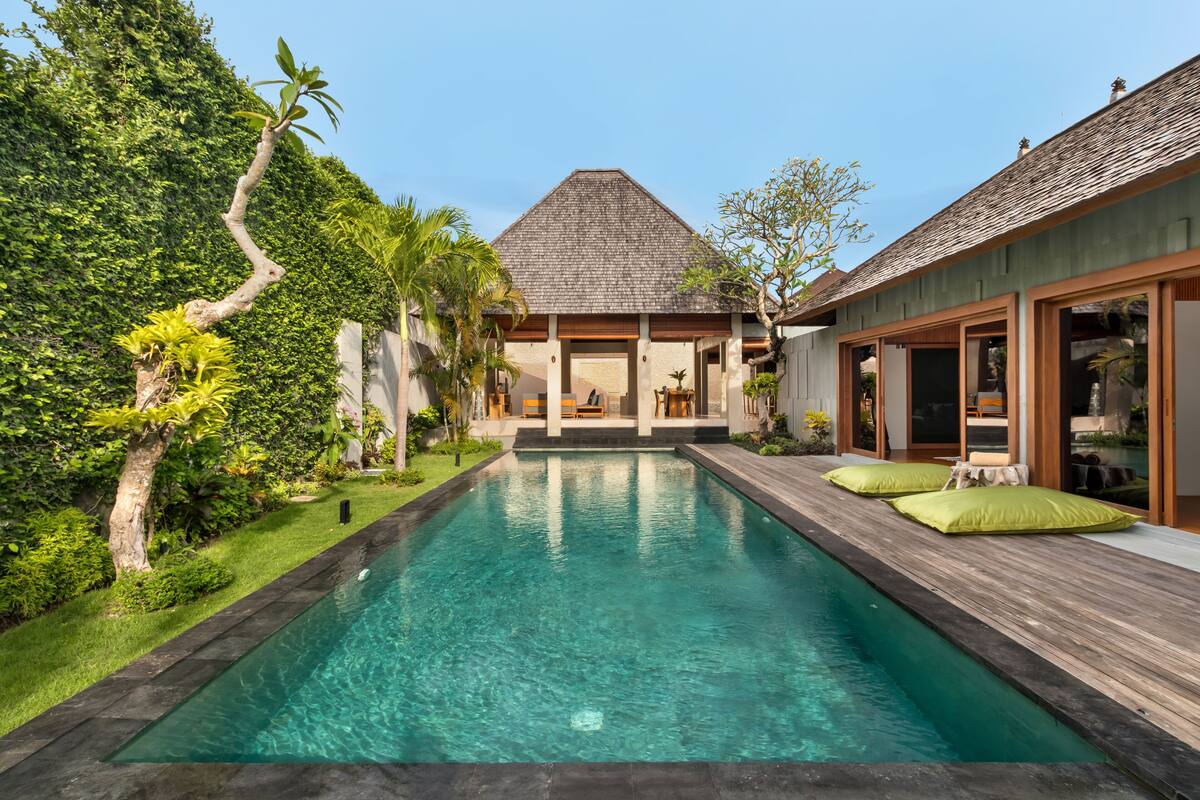 A pool with a wooden deck, surrounded by lush greenery and a house with a tiled roof.