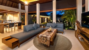 A modern living room with a sofa, a coffee table, and a TV. There is a dining area with a table and chairs. The room has large windows and a view of the outdoors. - Navya Villas (Kerobokan)