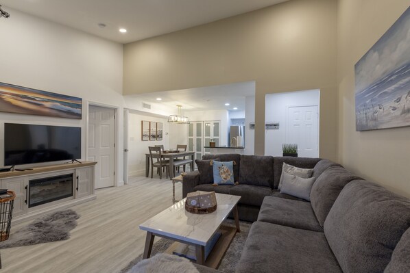 A modern living room with a grey sofa, a flat-screen TV, and a dining area with a chandelier.