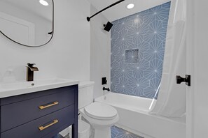 Casa de banho moderna com painel de azulejos geométricos, sanita branca, banheira e toucador com puxadores dourados.