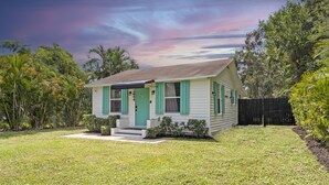 Une maison de plain-pied avec une porte et des volets verts, entourée d’une pelouse bien entretenue et de diverses plantes.