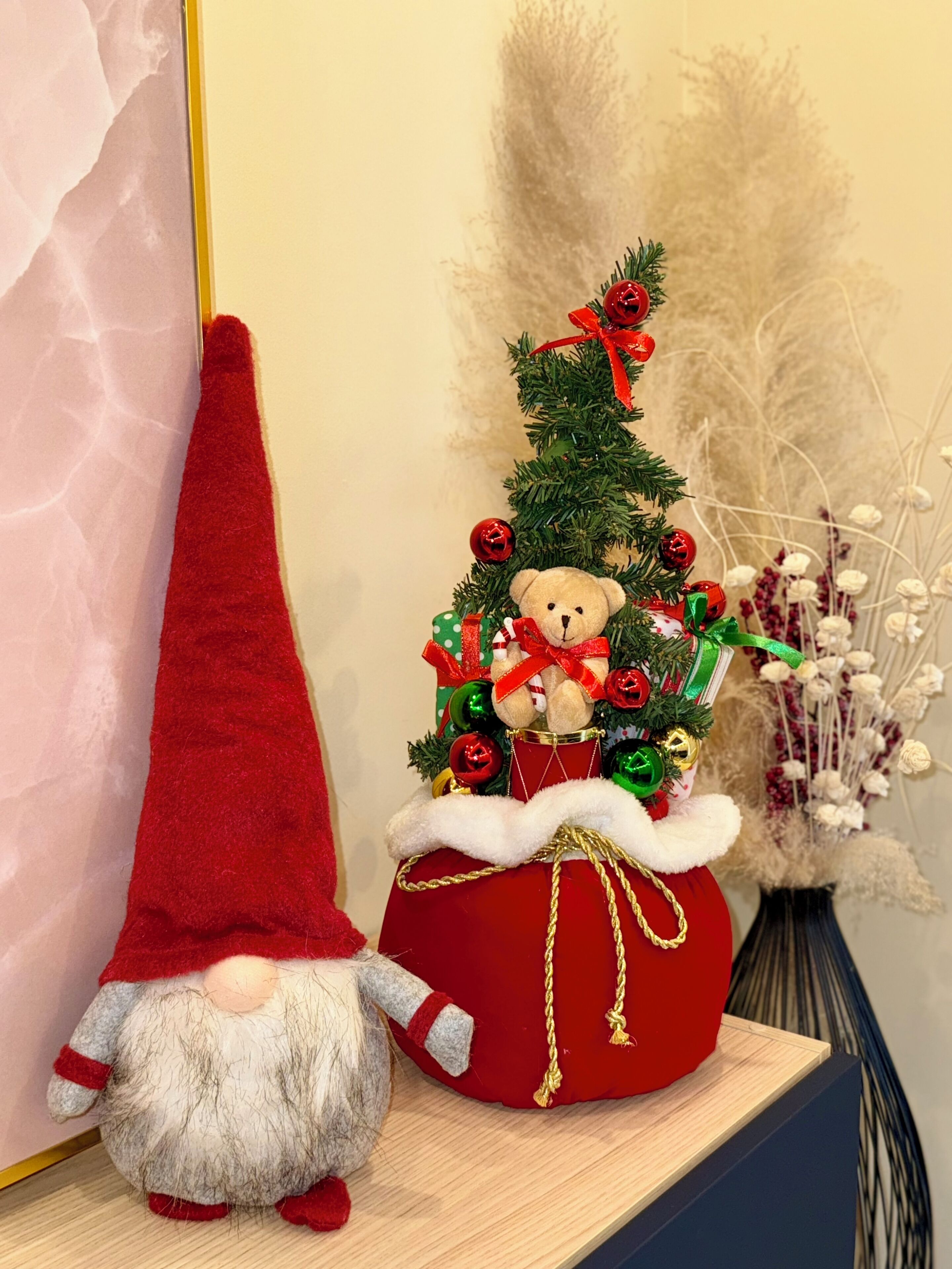 A Christmas tree with a teddy bear and a gnome, a red cone, and a vase with dried flowers.
