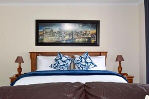 A hotel room with a bed, two nightstands, a framed cityscape painting, and a view of a river.
