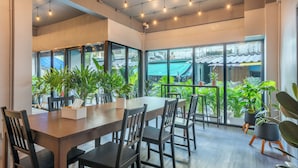 A spacious dining area with a long wooden table, multiple chairs, and potted plants. Large windows provide a view of the outside. - Ti Thong Hostel (Bangkok)
