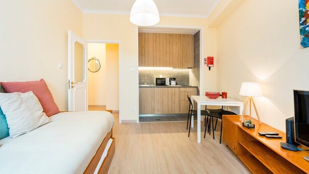 Habitación de hotel compacta con cama, mesita de noche, lámpara, televisor y una kitchenette con microondas y lavabo.