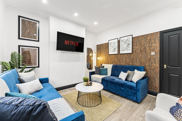 A modern living room with a blue sofa, a white coffee table, and a flat-screen TV mounted on the wall. - Stylish Designer 4 bed with Free Parking (Leeds)