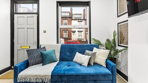 A blue sofa with patterned cushions, a white door, and a window with a view of buildings and a red car.