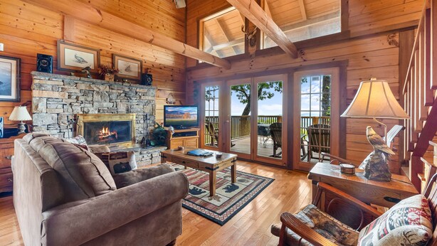 A cozy log cabin interior with a stone fireplace, comfortable seating, and large windows offering views of the outdoors. - Resort*Panoramic Views*Gameroom*Gas Grill*Hot Tub*Wifi/Cable (GATLINBURG)