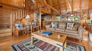 A cozy log cabin interior with a wooden staircase, a living room with a brown leather sofa, a dining area, and a kitchen. - Resort*Panoramic Views*Gameroom*Gas Grill*Hot Tub*Wifi/Cable (GATLINBURG)