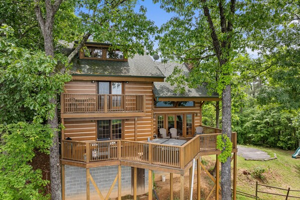 A multi-level wooden cabin with a deck, surrounded by trees. - Resort*Panoramic Views*Gameroom*Gas Grill*Hot Tub*Wifi/Cable (GATLINBURG)
