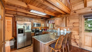 A wooden cabin kitchen with a central island, stainless steel appliances, and a dining area with wooden chairs. - Resort*Panoramic Views*Gameroom*Gas Grill*Hot Tub*Wifi/Cable (GATLINBURG)