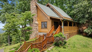 A log cabin with a stone chimney, wooden deck, and a view of the surrounding forest. - Resort*Panoramic Views*Gameroom*Gas Grill*Hot Tub*Wifi/Cable (GATLINBURG)