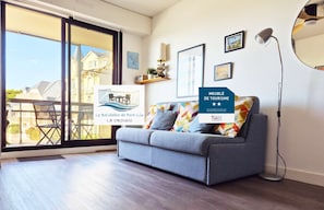 A modern living room with a grey sofa, colorful cushions, a floor lamp, and a large window with a view of buildings outside.