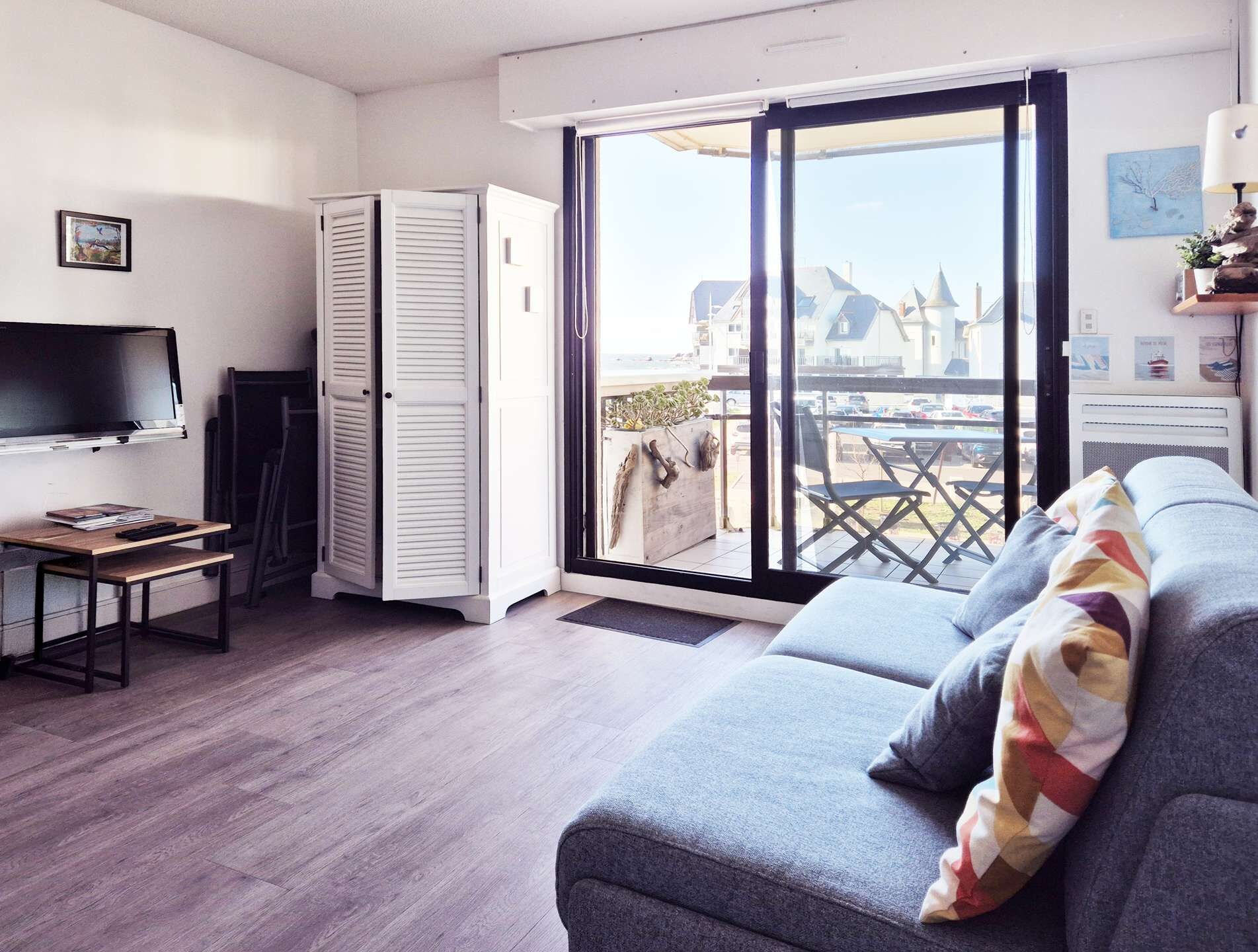A modern living room with a grey sofa, a flat-screen TV, and a sliding glass door leading to a balcony.