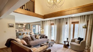 A spacious living room with a brown sofa, a beige armchair, and a coffee table. There is a kitchen in the background with a bar and stools. - Secret Room-Hot Tub-Billards-Firepit-Games-Pool access (Gal)