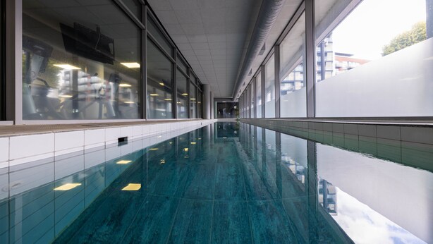 A long, narrow indoor swimming pool with clear glass walls reflecting the surroundings.